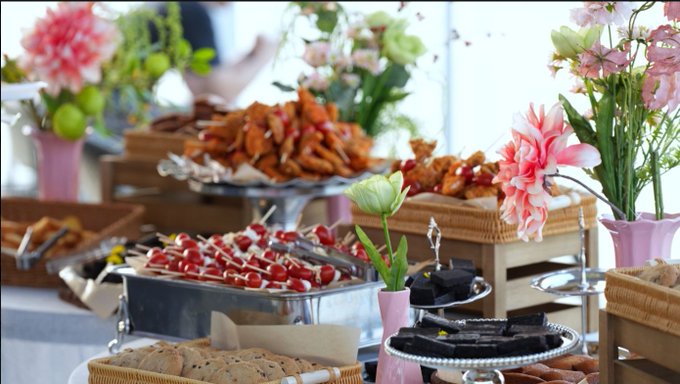 A catering buffet spread features skewers of meat and vegetables in metal containers, alongside pastries and pink carnations in small vases.