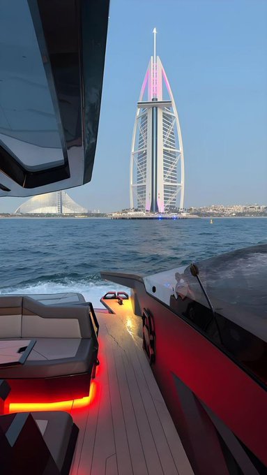A sleek red and black boat with illuminated interior flooring speeds across the water toward the Burj Al Arab hotel in Dubai, which is visible in the background.
