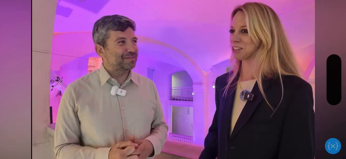 The image shows a man and woman standing next to each other with their hands clasped together, possibly having a discussion or posing for a photo. The background of the image features an archway that has purple lights shining through it, creating a unique atmosphere in the room where they are located.
