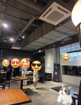 An indoor lounge or event space with dark ceilings and exposed ductwork features tables, a projection screen displaying a website, and decorative emoji stickers (heart-eyes and sunglasses faces) placed over a person in the center of the room.