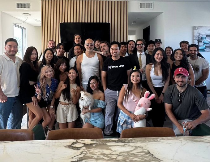 A diverse group of around 25 young adults pose for a group photo in a modern living room, with one man in the center wearing a "RYZE" branded shirt. A small white dog and someone holding a pink stuffed bunny add a playful touch to the gathering, supporting the caption's observation that content creator parties tend to have notably more women than typical web3 events.
