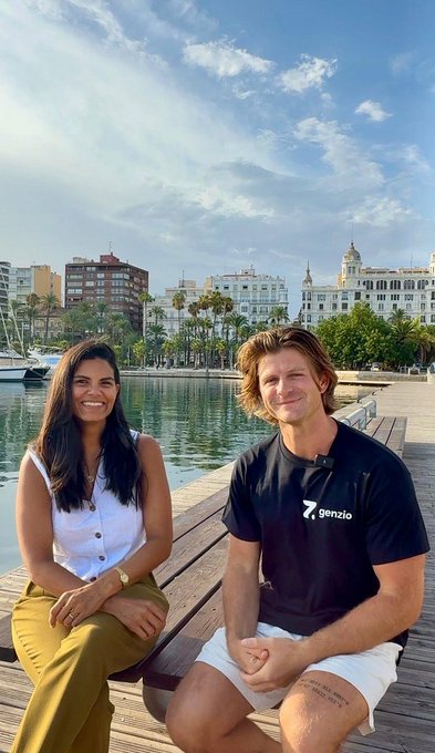 A woman in a white top and a man wearing a 'genzio' t-shirt sitting together by a waterfront with boats and buildings in the background.