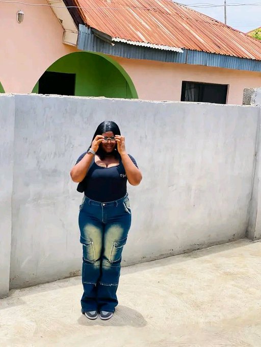 A photo of a woman standing outdoors against a gray concrete wall, wearing a dark blue top and faded blue jeans, holding a small object near her face.