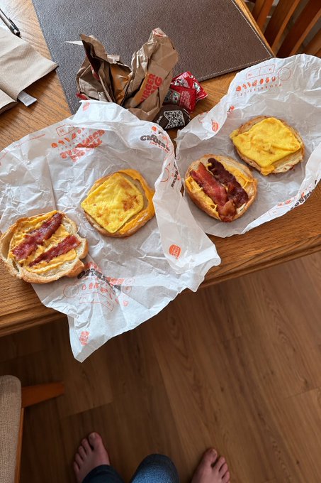 A spread of four open-faced breakfast sandwiches—two with bacon and egg, two with melted cheese—rests on Chick-fil-A wrappers atop a wooden table, with a person's bare feet visible on the floor below. The post appears to celebrate enjoying the meal while sharing a Solana (CA) wallet address for further engagement.
