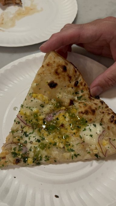 A hand reaches toward a triangular slice of pizza topped with herbs, corn, red onions, and green sauce on a white paper plate.