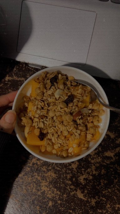 A photograph of a hand holding a white bowl filled with a mango dessert topped with granola and other ingredients, with a spoon resting inside, sitting on a dark countertop.