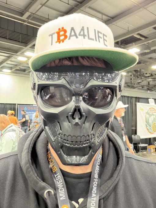 A person at a convention wearing a metallic skull mask, dark sunglasses, and a white cap with the Bitcoin logo and the text 'TBA4LIFE'.