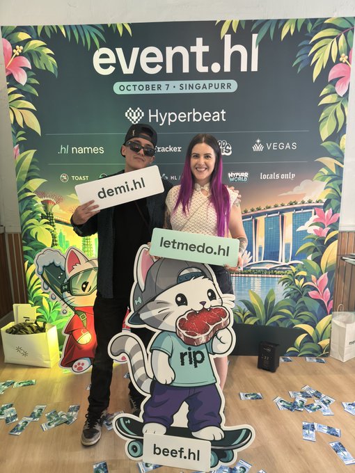 Two people pose at an event.hl photo wall dated OCTOBER 7 · SINGAPURRE, holding signs 'demi.hl' and 'letmedo.hl' in front of a cartoon cat mascot holding a meat 'rip' sign labeled 'beef.hl', with logos including Hyperbeat and VEGAS.