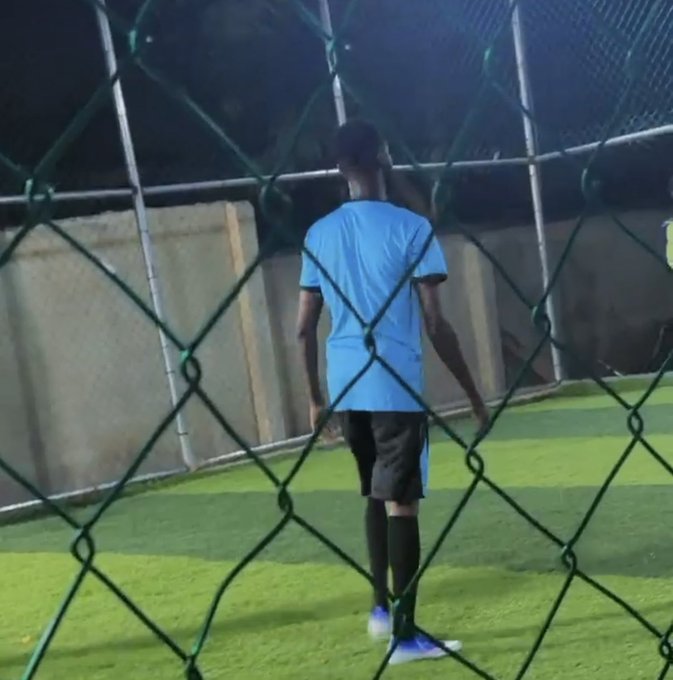 A person in a light blue soccer jersey, black shorts, and blue shoes stands on a green turf field behind a chain-link fence, appearing to be playing or practicing soccer.