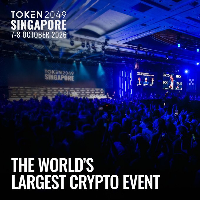 A packed conference hall bathed in blue lighting with a stage screen displaying "TOKEN2049 SINGAPORE," promoting the world's largest crypto event taking place October 7-8, 2026 at Marina Bay Sands with 25,000+ attendees, 300+ speakers, and 500+ exhibitors, with early-bird tickets at $399 rising to $499 on Friday.