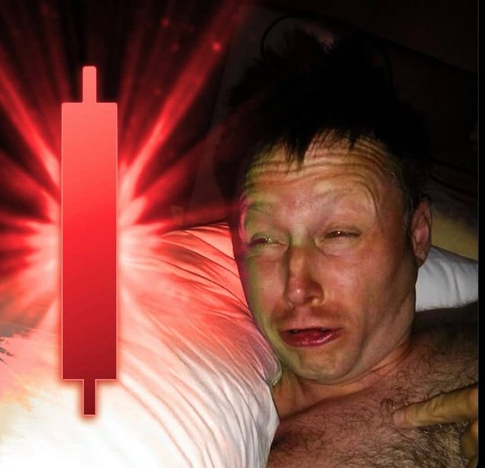 A selfie of a shirtless man lying in bed with a distressed expression, overlaid with a large red downward-trending candlestick chart graphic on the left side.