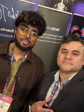 Two men pose for a selfie at what appears to be a convention, wearing lanyards with event badges against a backdrop of sponsor logos.