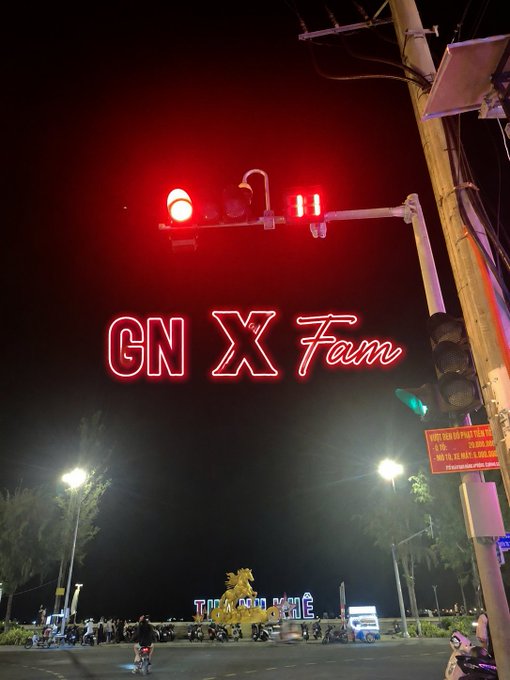 A nighttime photograph of a traffic light glowing red, overlaid with neon-style text reading 'GN X Fam', with a golden statue and crowd visible in the background.