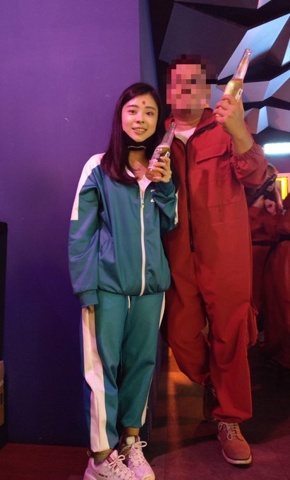 Two people dressed in teal and red tracksuits stand together holding bottles, resembling characters from Squid Game, in a room with purple lighting.