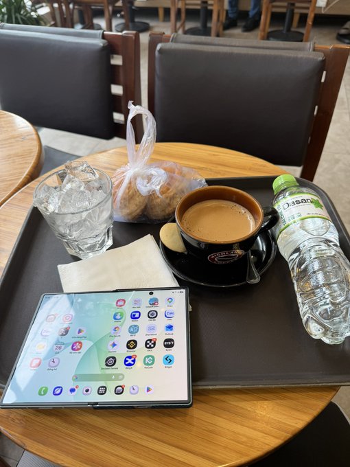 A wooden cafe table holds a tray with a cup of coffee, a glass of ice water, a bottle of Pocari Sweat, a pastry, and an unfolded Samsung Galaxy Z Fold phone displaying its home screen.