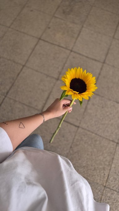 A person with a triangle tattoo on their forearm holds a sunflower over a tiled floor, wearing a white skirt and silver bracelet.