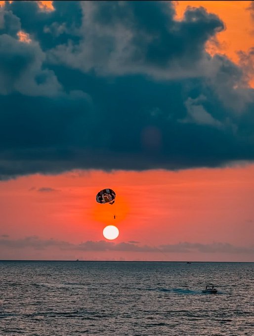 A scenic sunset photograph over the ocean featuring a bright orange sun low on the horizon beneath dramatic dark clouds, with a parasailer floating in the sky and a small boat on the water below.