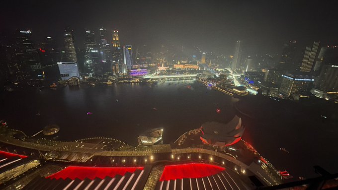A nighttime aerial view of Singapore's Marina Bay waterfront, captured from the rooftop of Marina Bay Sands with its iconic illuminated red structures in the foreground, overlooking the city skyline and harbor. The image reflects the "c'est la vie" sentiment of unwinding following Token2049, showcasing the glamorous, high-altitude Singapore setting typical of post-conference nights in the city.