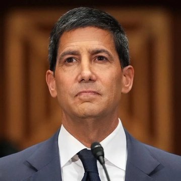 A serious-looking man in a dark suit and tie sits before a microphone, appearing to be at a formal hearing or press conference. The bullish caption "SEND ALL OUR COINS" suggests the image is being used to imply a positive market sentiment or call to action, possibly referencing the individual pictured in a financial context.