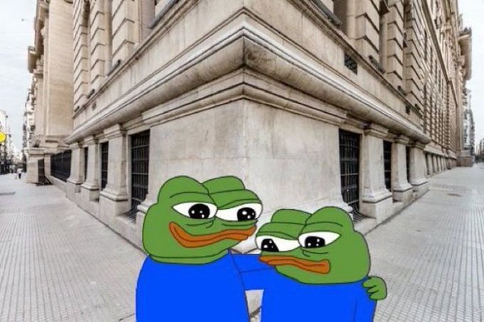 Two cartoon Pepe the Frog characters in blue shirts hug each other while standing on a city street corner in front of a large stone building.