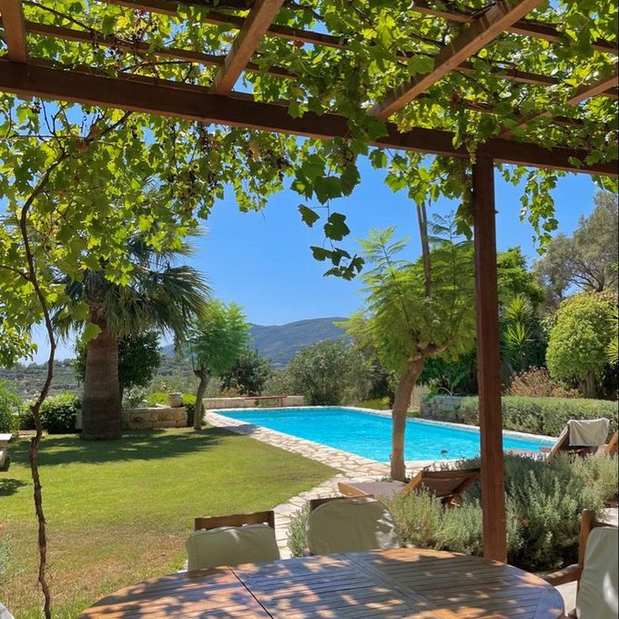 A serene countryside retreat in Italy featuring a pergola draped in lush grapevines, overlooking a turquoise pool surrounded by manicured gardens and rolling hills. The scene perfectly captures the post's vision of escaping to a peaceful Italian villa with land to enjoy the fruits of crypto success.