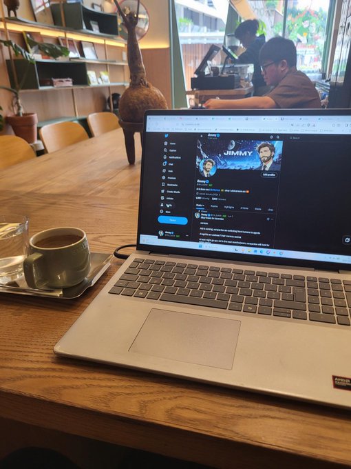 A laptop on a wooden café table displays Twitter/X with a 'JIMMY' profile, alongside a ceramic coffee cup, with patrons and plants visible in the background.