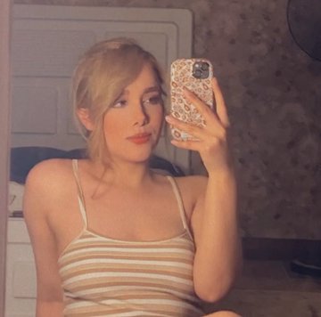 A young blonde woman in a striped tank top takes a mirror selfie in a bedroom, holding a phone with a patterned case.