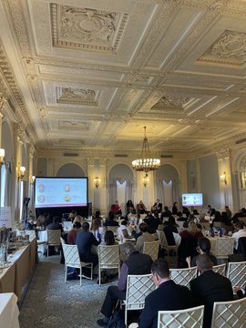 A crowded conference or presentation taking place in an ornate, classical-style room with a decorative coffered ceiling and chandelier. Attendees sit at tables facing a speaker and projection screens displaying charts.