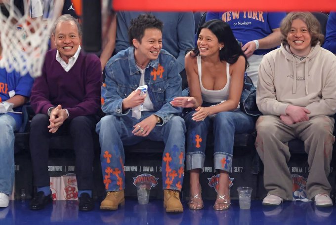 The image shows a group of people sitting together in front of a basketball court. There are four individuals visible - two men and two women. The man on the left side of the image has his arms crossed, while the woman next to him is smiling at something or someone. Another person can be seen seated further back in the scene.

The group appears to be enjoying their time together, possibly watching a basketball game or engaging in conversation. There are two cups placed near them on the bench they're sitting on, suggesting that they might have been drinking water during breaks in the game.