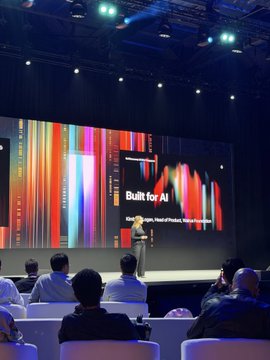 A conference presentation with a speaker on stage displaying a slide titled "Built for AI" with colorful graphic designs. Audience members are seated in chairs watching the presentation.