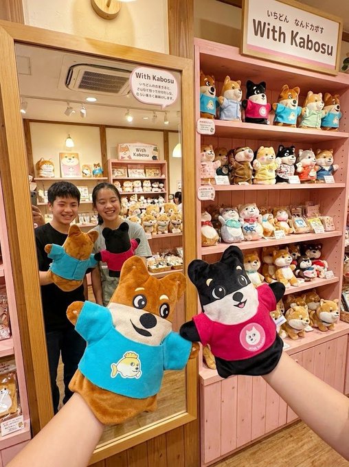 Two hand puppets of Shiba Inus wearing blue and pink shirts are held up in front of a mirror inside the "With Kabosu" store, which displays shelves of plush toys and merch featuring the iconic Doge dog. The auction raised 5169 $DOGE and $475 to support animal welfare causes.