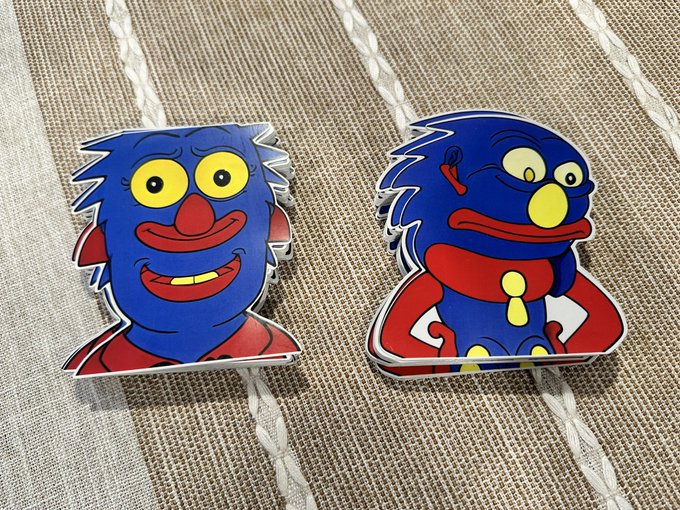 Two rubber coasters or magnets featuring blue, red, and yellow cartoon monster designs with spiky hair and wide smiles, sitting on a woven tan surface with white braided trim.