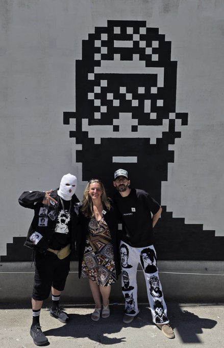 The image features three people posing for a photo next to a large black and white graffiti mural of a man's face, which appears to be a mask or a character from a video game. The group consists of two men wearing masks and one woman without any mask. They are standing in front of the mural on a sidewalk, with the background being a wall.

The caption for this image reads: "Only a few understand Proud Normie Love this community Much". This phrase suggests that the people posing for the photo might be fans or admirers of the character depicted in the graffiti mural, and they are likely discussing their shared interest.