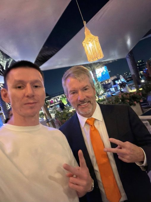 The image shows a man wearing an orange tie standing next to another man who has his hands in the air, making peace signs. Both men appear to be wearing suits and ties, suggesting they are dressed for a formal event or occasion. The backdrop of their gathering is a white tent with lights inside it, indicating that the photo was taken at night. There seems to be some confusion surrounding why a man's $2,500,000 worth of bitcoin (BTC) should pay for his income from the sale of another $2,000,000 worth of Bitcoin on February 28th, as TF has set up an automated system to do this. The image is captioned "Once again why TF is @saylor trying to fund a dividend with $2,500,000 by sale ling $btc ? Why didn't he just set aside $2,500,000 from the almost $2,000,000,000 that he spent to buy Bitcoin to do that? Something is not adding up".