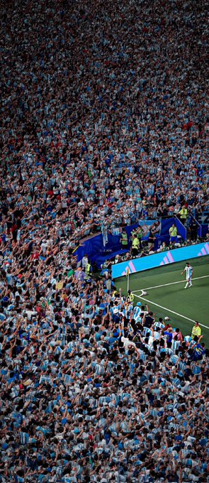 A massive crowd of Argentina soccer fans in light blue and white jerseys fills a stadium section, cheering from the stands next to the corner flag of a soccer field.