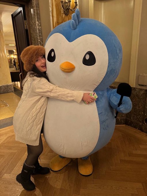 A woman wearing a furry winter hat and cream sweater playfully hugs a large blue and white penguin mascot indoors.