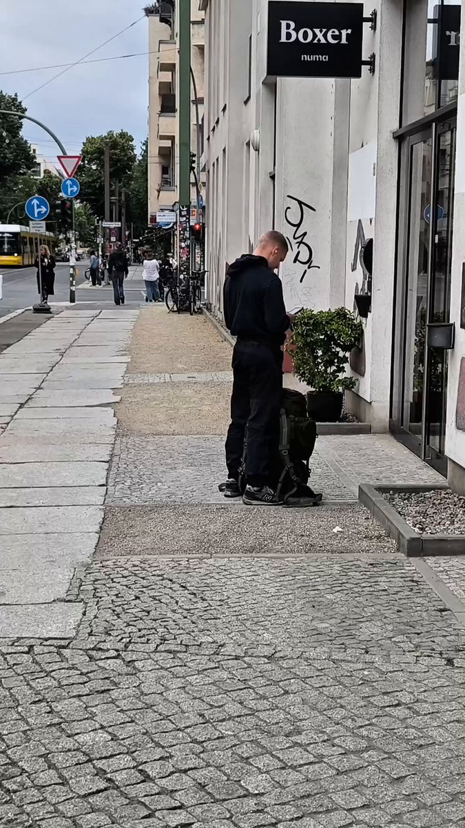 The image shows a man standing outside of a store, possibly a clothing or shoe shop, wearing black clothes and holding a backpack. The man appears to be looking at something inside the store window. There are several other people visible in the background, some walking on the sidewalk and others standing near parked cars. A bus is also present in the scene, indicating that this location might be close to public transportation.