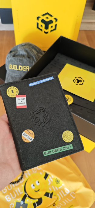 A black leather notebook labeled 'BUILDER' with various crypto-themed stickers including 'HEZZ BULLISH' and 'BUILDERS ONLY', held in front of a yellow BNB-branded gift bag and merchandise.