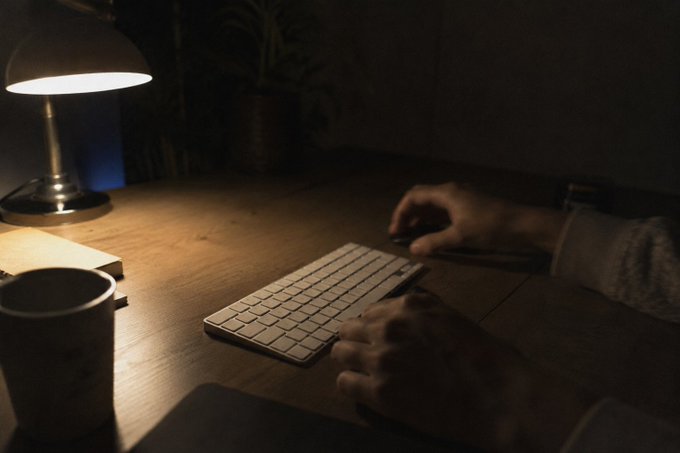 The image shows a person sitting at a desk with a keyboard in front of them. The person's hands are positioned over the keys, suggesting they might be typing or using an external device connected to the keyboard. There is also a cup on the desk next to the keyboard, possibly containing coffee or another beverage for the person working at their computer.