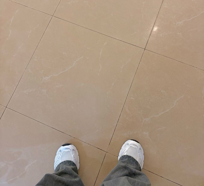 A first-person view looking down at white sneakers and gray pants standing on beige marble floor tiles.