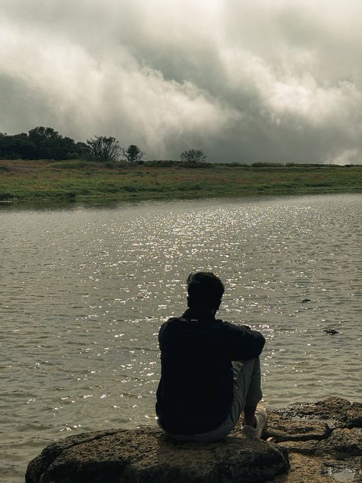 A person sits in silhouette on a rock by a shimmering lake, facing a cloudy, overcast sky and distant tree-lined shore.