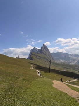 A scenic mountain landscape on a sunny day, showing hikers walking along a dirt trail through a green alpine meadow toward a rocky peak.