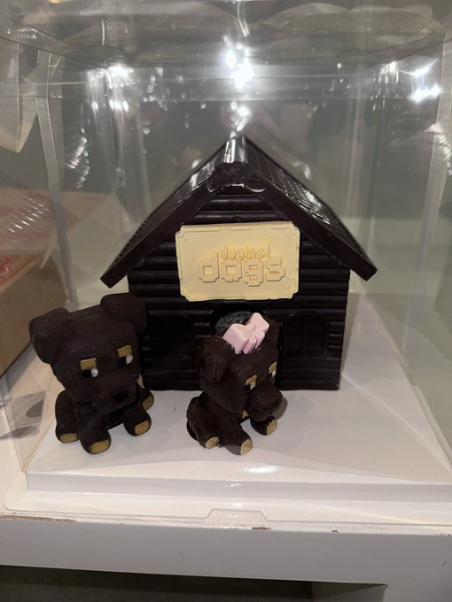 A chocolate sculpture displayed in a clear case features a dog house with the 'Boston dogs' logo, accompanied by two small chocolate dog figures.