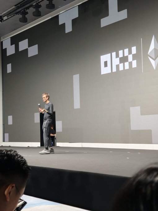 A man stands on a stage holding a microphone in front of a large screen displaying "OKX" and the Ethereum logo, with pixelated graphics in the background and an audience visible in the foreground.