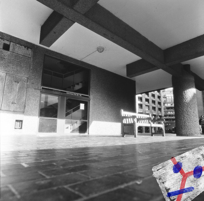 A black-and-white photo shows an empty bench tucked beneath the concrete overhang of a Brutalist building. A small sticker with a blue figure sits on the tiled floor in the foreground, echoing the post's "September = off the bench" line about finally stepping out of retirement.