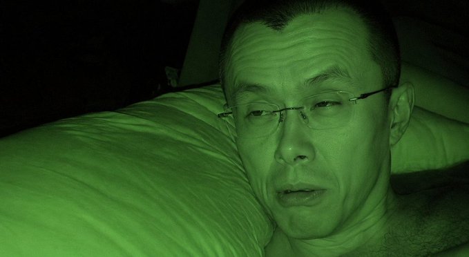 A close-up selfie of a man wearing rimless glasses lying down on a pillow, illuminated by green-tinted lighting in a dark room.