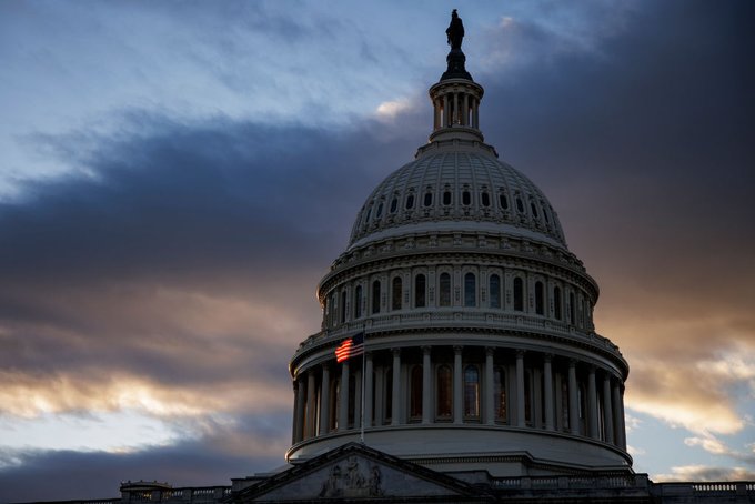 The United States Capitol building dome is captured at sunset against a dramatic sky with dark clouds and golden light, with an American flag visible on the side of the structure.