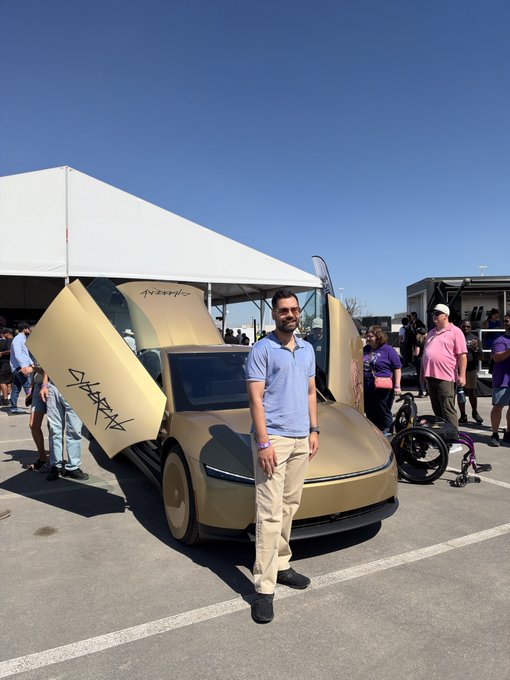 A man stands proudly in front of a gold-colored Tesla Cybertruck with graffiti on its doors, displayed under a tent at what appears to be an outdoor event.