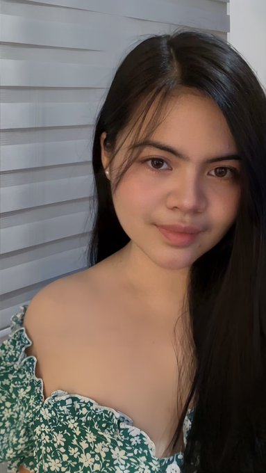 A young woman with long dark hair poses for a close-up selfie, wearing a green floral off-the-shoulder top against a background of white window blinds.