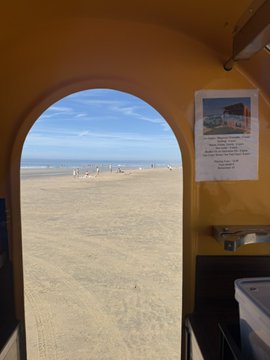 The image shows a beach scene with people enjoying their time near the water, some of them standing or walking along the shoreline. There are at least 12 visible individuals scattered across the sandy beach area. The archway leading into the photo provides an interesting perspective on the beach and its surroundings.